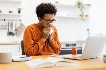 Obraz premium An African-American man wearing glasses sneezes into a tissue while working on a laptop at home.