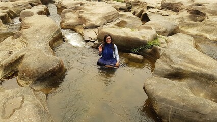 Serene woman embraces natural beauty, performing namaste gesture amidst tranquil rocky river landscape during peaceful travel adventure