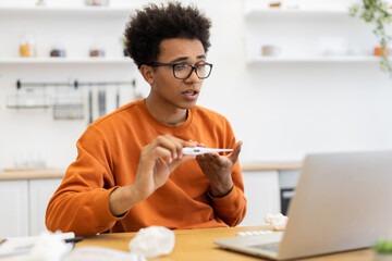 A young man with a thermometer is on a video call, possibly discussing his health while working from home.