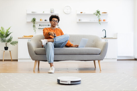 A young man watches TV while a robot vacuum cleaner cleans the floor in his modern living room.