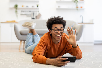 A young man lies on the floor, waving during a video call on his smartphone, smiling with joy.