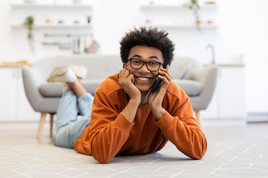 A young man with glasses lies on the floor, smiling while talking on his phone in a cozy living room setting.
