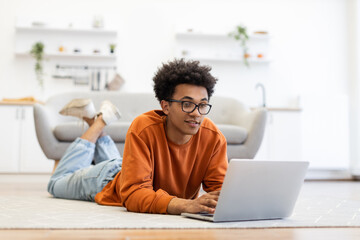 A young man with glasses is lying on the floor, using a laptop in a modern living room setting.