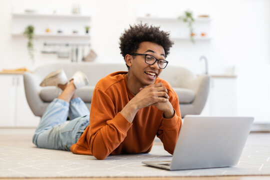 A young man with glasses lies on the floor, smiling while video conferencing on his laptop in a cozy home setting.
