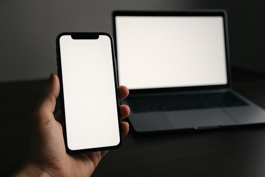 Hand holding smartphone displaying blank screen with laptop in background, symbolizing technology, connectivity, and digital communication concepts.