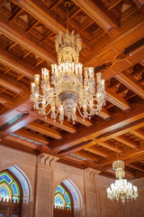 A luxurious interior featuring grand crystal chandeliers hanging from an intricately carved wooden ceiling. The architectural design includes detailed Islamic patterns, arched windows
