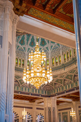 A magnificent interior view of an Islamic mosque featuring a large golden chandelier suspended beneath an intricately patterned turquoise dome. The space is adorned with detailed geometric designs