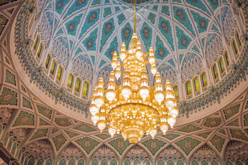 A interior view of an ornate Islamic dome featuring intricate geometric patterns, turquoise mosaics, and a massive golden chandelier. The architectural beauty and craftsmanship of Middle East design