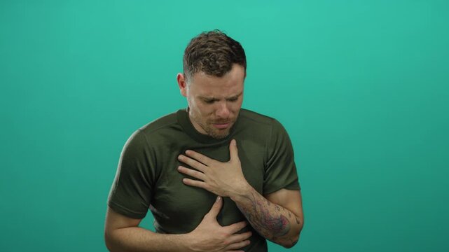 Young man with tattoos in distress wearing green shirt against isolated green background gripping stomach showing emotional struggle and concern