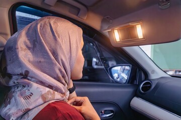 A Muslim woman looking in the mirror on car sun visor while adjusting her hijab in a car