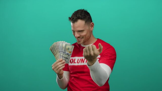 Young man holding american dollars in a red shirt with volunteer text on isolated green background displaying wealth and generosity joyfully pointing and gesturing index finger.