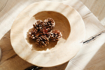 Pine cones resting in a simple wooden bowl on a textured wood surface with soft sunlight