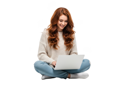 A young woman with long auburn hair sits cross legged on the floor with a laptop smiling and looking down at the screen isolated on transparent background