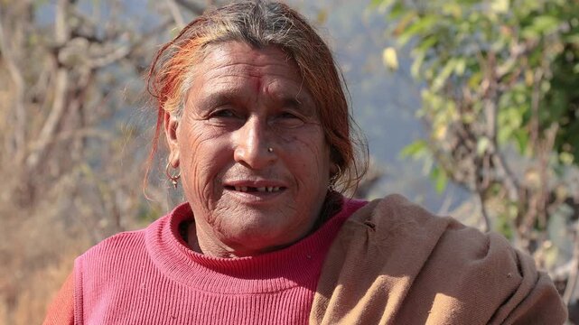 Close-up 4K footage of an elderly rural woman aged around 80 to 85 years, smiling outdoors in a peaceful mountain village of Uttarakhand, India, captured in natural sunlight with trees and hills in th