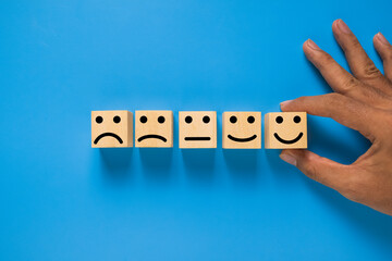 male hand put wooden blocks with five different emoji expression from sad to happy isolated on blue background. rating scale satisfaction feedback concept