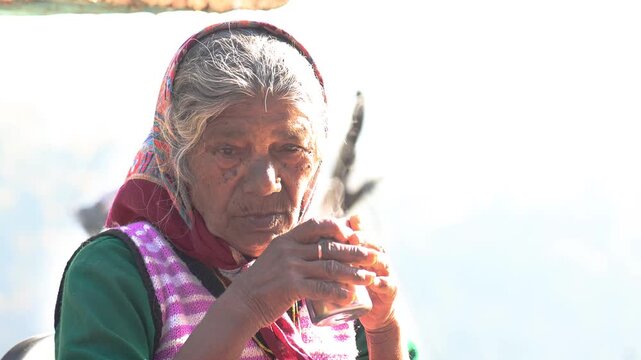 Old Indian rural woman around 80 to 85 years enjoying a warm cup of tea outdoors at her village home, sitting peacefully in a natural mountain setting.