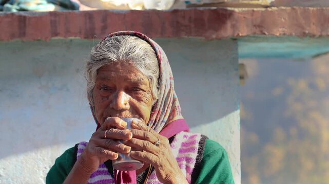 Old Indian rural woman around 80 to 85 years enjoying a warm cup of tea outdoors at her village home, sitting peacefully in a natural mountain setting.