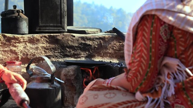 A traditional Indian village woman cooks roti on a clay chulha, gently flipping it on a hot iron tawa above an open flame. Her bangles shimmer in the sunlight as she prepares the meal with skill.