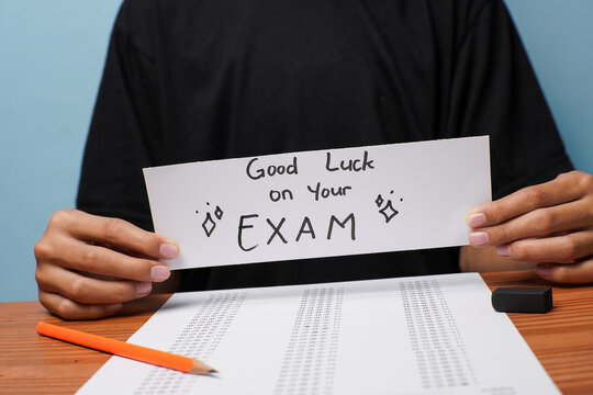 A student holding a handwritten good luck exam card centered in the frame above a test sheet and pencil, representing study motivation and exam preparation.