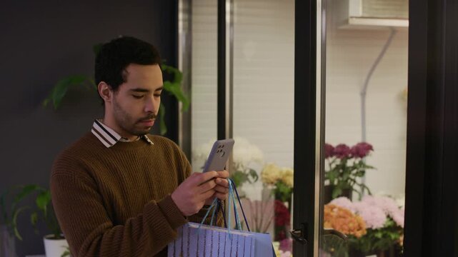 Medium shot of young male customer taking photo of bouquets on mobile phone in front of shop window while visiting florist shop