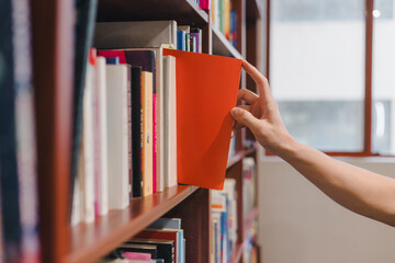 Hand picking a red book from library bookshelf in soft natural light. Ideal for concepts of education, knowledge, research, learning academic environments.