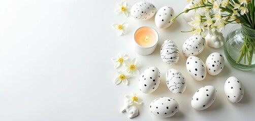 White Easter eggs with black speckled designs, fresh spring flowers, softly lit candle arranged flat lay. Glass vase holds white blooms, next to small ceramic bunny figurine. Decor creates modern,