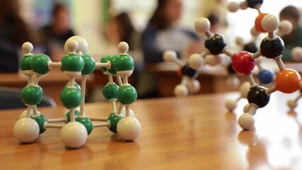 Two molecular models with green, white, black, orange and blue spheres are on a wooden table in a classroom with blurred students, representing science education. - Powered by Adobe