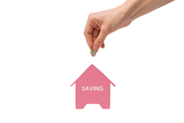 Hand drops a coin into a piggy bank house. Focus on financial saving, down payment, and home ownership goals.