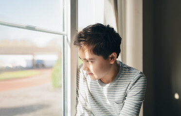 Young boy looking out the window with natural sunlight, expressing emotions of loneliness, daydreaming, or hope. Captures peaceful and introspective mood of thoughtful Teen boy