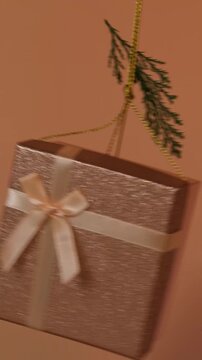 A beige shiny gift box swings from side to side like a pendulum with a spruce branch on a shiny gift rope on a beige background