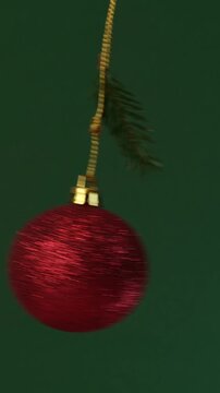 A red shiny Christmas ball swings from side to side like a pendulum with a spruce branch on a rope on a green background
