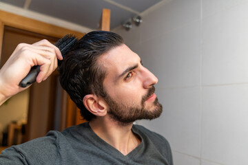 Obraz premium Young man holding a brush, styling his dark hair and beard, practicing his morning beauty and personal care routine