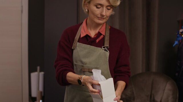 Tilt up shot of adult Caucasian woman in apron wrapping vase in paper and placing in small box at table in gift shop