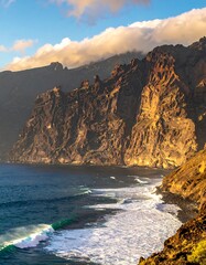 A coastal scene showcases rugged cliffs meeting ocean. Sunlight bathes the rock face, while waves crash against the shore. Clouds