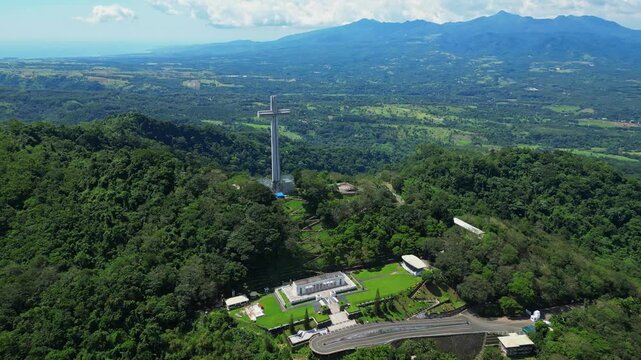 Left pan aerial showcasing the towering Mt. Samat National Shrine, revealing its towering cross monument framed by lush forested hills and sweeping valleys in Pilar, Bataan, Philippines.