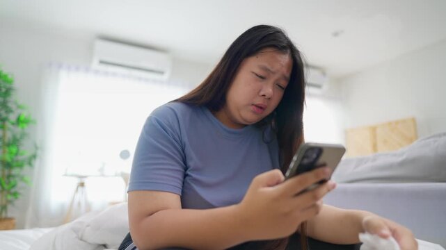 Unwell Asian young plus-size woman using smartphone in cozy room at home. 