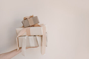 Birthday, Christmas gift concept. Female hand holding stack of gift boxes with beige ribbons over white wall. Holiday background with blank copy space