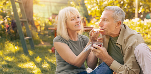 A happy couple sits on the grass in a garden during a sunny afternoon. The woman offers a slice of apple to the man, who smiles in delight. They enjoy each other's company in a peaceful setting.
