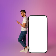 A young Arab man is engaged with his mobile device while leaning against a large smartphone with a blank screen.