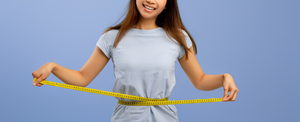 A young woman poses cheerfully while measuring her waist with a tape measure against a blue background. She is dressed in a casual gray shirt, embodying positivity and health-consciousness.
