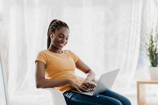 A young black businesswoman in earphones smiles while having a video call. She is working from her home office, engaging with colleagues during an online meeting.