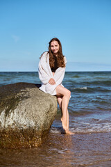 Young woman sitting on rock by Baltic Sea shore under clear blue sky