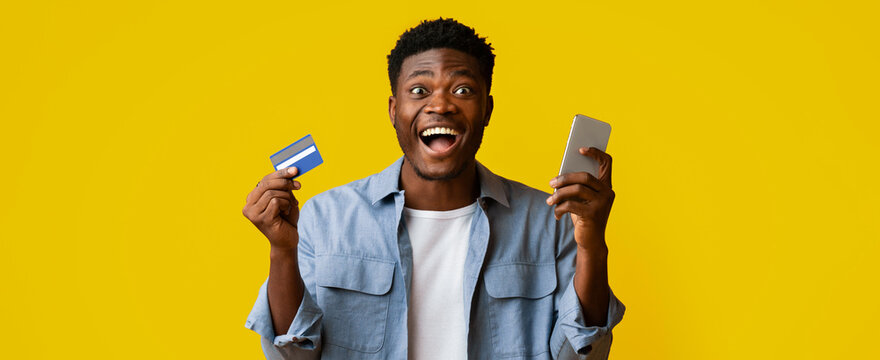 A man is expressing joy while holding a credit card in one hand and a smartphone in the other. He stands against a vibrant yellow background, showcasing excitement and positivity.