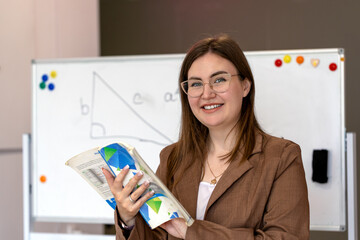 Confident young woman in glasses holding a folder while standing in front of a whiteboard. Professional teacher or businesswoman in a classroom or office setting