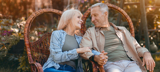 A couple sits closely together in a cozy outdoor chair, smiling and holding hands. They enjoy a serene moment surrounded by flowers and greenery, radiating warmth and love.