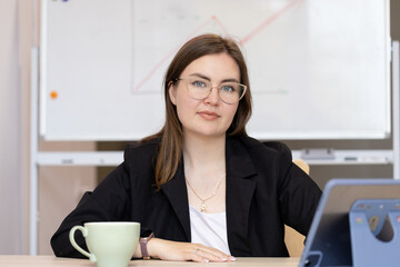 Confident young woman in glasses holding a folder while standing in front of a whiteboard. Professional teacher or businesswoman in a classroom or office setting