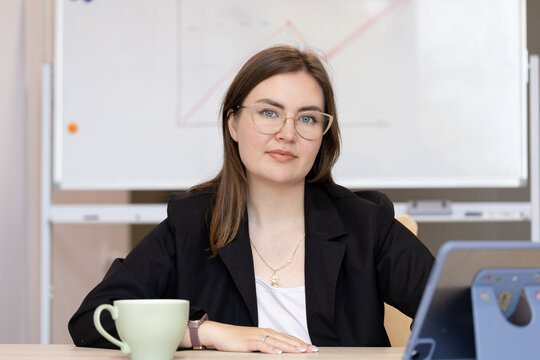 Confident young woman in glasses holding a folder while standing in front of a whiteboard. Professional teacher or businesswoman in a classroom or office setting