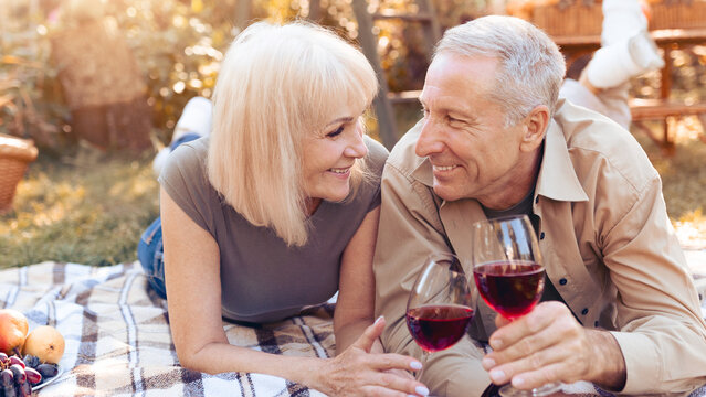 A happy couple lies on a blanket in a garden, sharing a joyful moment over glasses of red wine. They smile at each other, surrounded by fruits and a peaceful outdoor setting.
