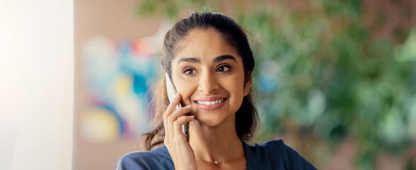 A young woman is smiling while holding a smartphone to her ear. She appears engaged in conversation, surrounded by greenery. The background features colorful artwork.