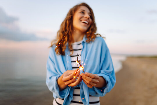 A joyful woman with curly hair stands on the shore holding sparklers at sunset. The beautiful woman enjoys the seascape and creates a festive atmosphere in nature. Concept of celebration and walks.